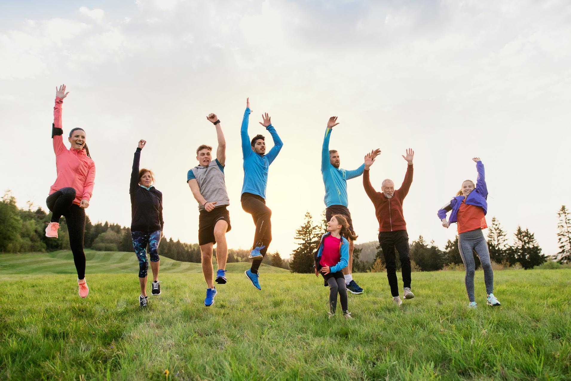 Gruppe von sieben Menschen springt fröhlich in einer Wiese, umgeben von Bäumen und einem hellen Himmel.