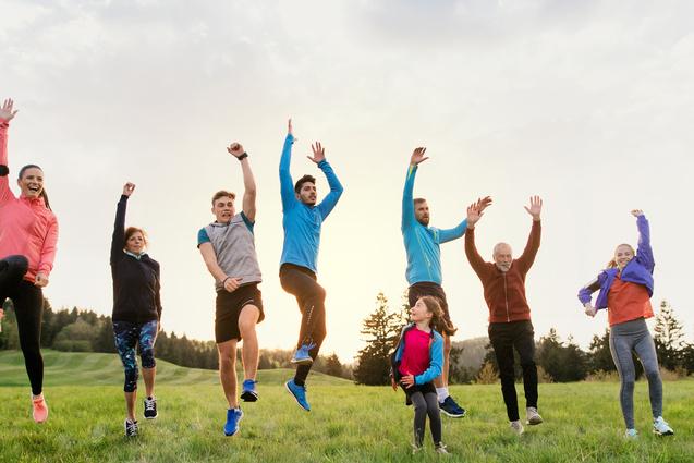 Gruppe von sieben Menschen springt fröhlich in einer Wiese, umgeben von Bäumen und einem hellen Himmel.