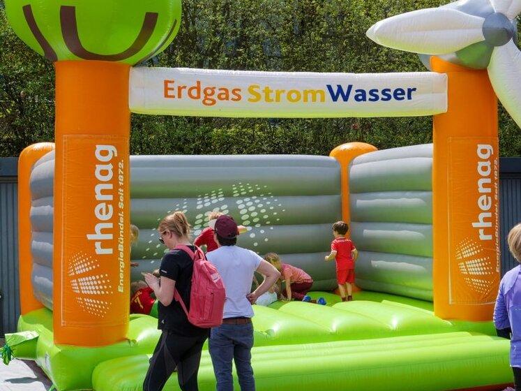 Eine Hüpfburg in den Fraben Orange, Weiß und Hellgrün vom Sponsor Rhenag. Die Linke Säule der Hüpfburg soll ein Baum darstellen und die rechte ein Windrad. Auf und neben der Hüpfburg sind Leute abgebildet.