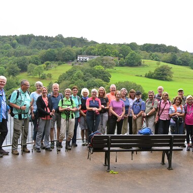 Gruppe von Wanderern steht auf Aussichtsplattform mit Blick auf einen See und grüne Hügel. Sitzbank im Vordergrund.