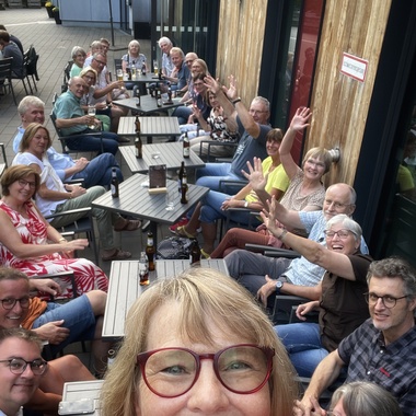 Gruppenaufnahme von Menschen an einem Tisch im Freien, alle lächeln und winken, mit Getränken auf dem Tisch.