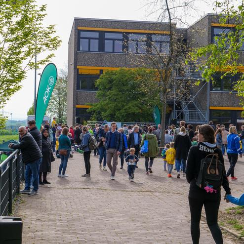 Menschenmenge bei einer Veranstaltung vor einem Gebäude, umgeben von Bäumen und AOK-Flaggen.