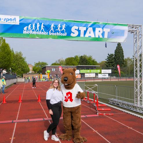 Frau in weißem Shirt steht neben einem kostümierten Bären an der Startlinie eines Lauf-Events auf einem Sportgelände.