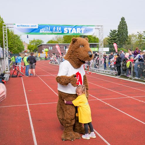 Ein Bärmaskottchen umarmt ein kleines Mädchen auf einer Laufbahn vor dem Startbanner einer Veranstaltung.