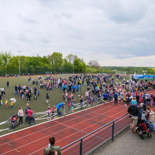 Große Menschenmenge auf einem Sportplatz, verteilt zwischen Spielständen und einer Ziellinie, umgeben von Bäumen.
