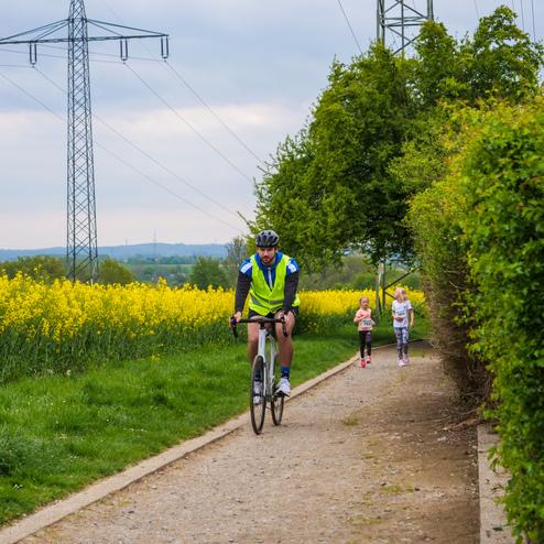 Radfahrer auf einem Weg, begleitet von zwei joggenden Kindern, mit gelben Rapsfeldern im Hintergrund.
