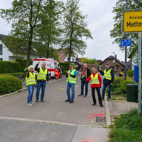 Gruppe von sechs Personen in Warnwesten stehen an einer Straße vor einem Ortsschild 'Kreisstadt Mettmann'.