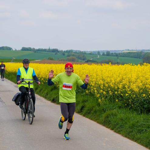 Läufer in einem grünen Shirt winkt auf einem Weg, flankiert von Rapsfeldern und Radfahrer in Hintergrund.