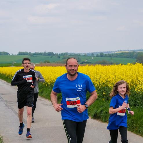 Läufer in blauen T-Shirts joggen auf einem Weg, umgeben von leuchtend gelben Rapsfeldern und grünen Hügeln.