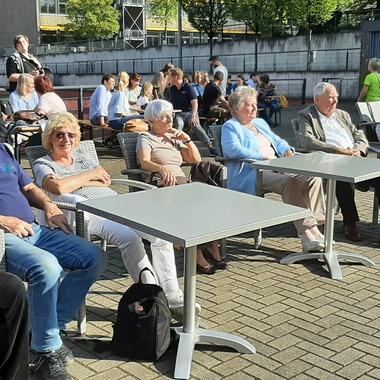 Gruppierung von älteren Menschen in Stühlen, die auf einer Terrasse vor einem Sportplatz sitzen.