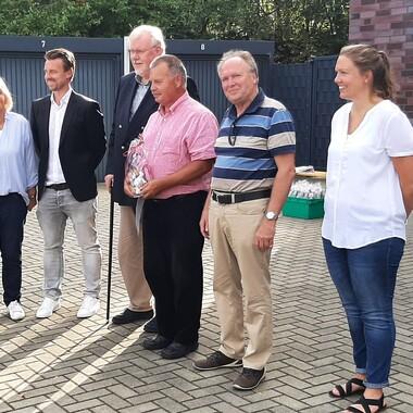 Sechs Personen stehen auf einer gepflasterten Fläche, einige lächeln; Hintergrund mit Garagentoren und Bäumen.