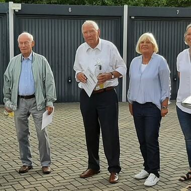 Fünf Personen stehen zusammen vor Garagen, einige halten Papiere und eine Flasche, im Hintergrund graue Wände.