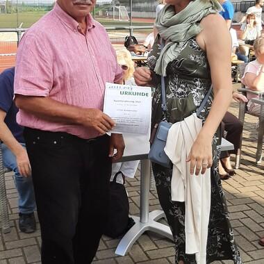 Mann mit Urkunde steht neben Frau, die lächelt; im Hintergrund Tische und ein Sportfeld sind sichtbar.