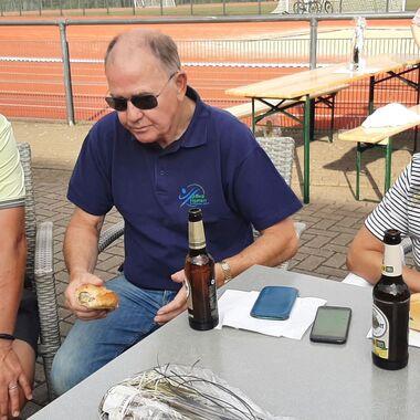Drei Personen sitzen an einem Tisch, mit Getränken und Snacks, im Hintergrund sind andere Menschen und ein Sportplatz sichtbar.