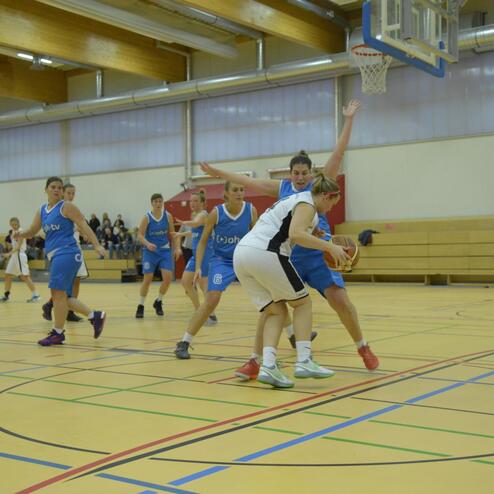 Spielszene in einer Basketballhalle: Spielerinnen in blauen und weißen Trikots kämpfen um den Ball auf dem Spielfeld.
