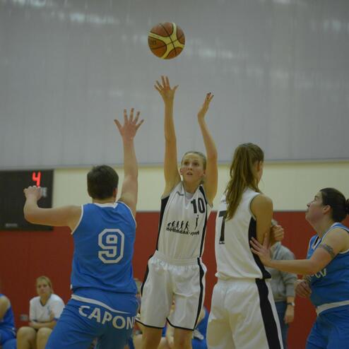 Spielerin in weißem Trikot wirft Ball während eines Basketballspiels, gegnerische Spielerinnen stehen in blauen Trikots.