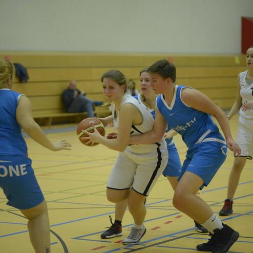 Zwei Basketballspielerinnen kämpfen um den Ball während eines Spiels in einer halben Sporthalle.