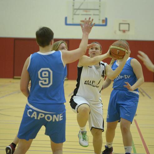 Basketballspiel: Spielerin in schwarzer Trikots zieht zum Wurf, umgeben von Gegnerinnen in blauen Trikots.
