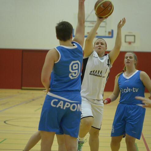 Basketballspiel: Spielerin in weißer Uniform wirft Ball über zwei Gegnerinnen in blauen Trikots auf einem Hallenboden.