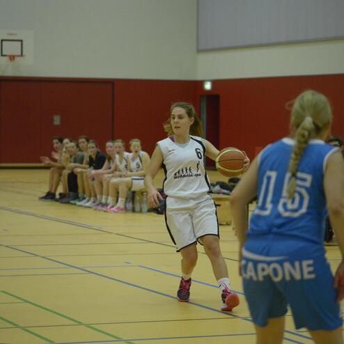 Spielerin in weiß dribbelt den Basketball auf dem Spielfeld, während die Gegnerin in Blau Position bezieht.