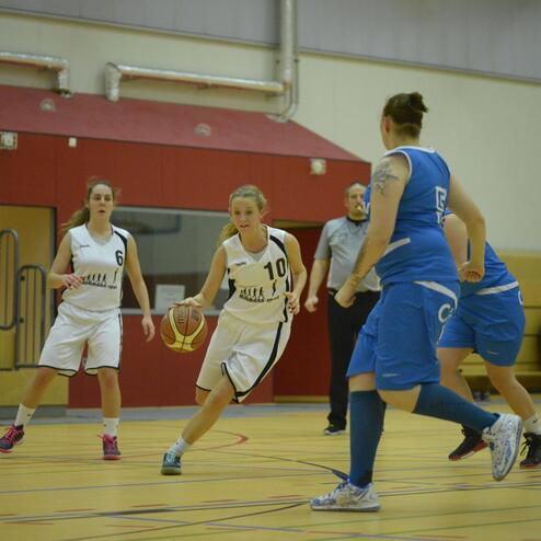 Basketballspiel mit zwei Teams: Spielerin in Weiß dribbelt den Ball, während Gegenspielerinnen in Blau reagieren. Zuschauer im Hintergrund.