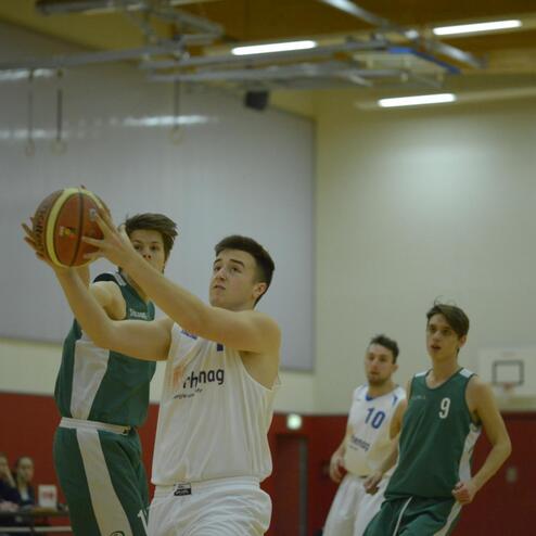 Spieler in weißen Trikots fängt einen Basketball, während andere Spieler in grünen Trikots auf einem Basketballfeld stehen.