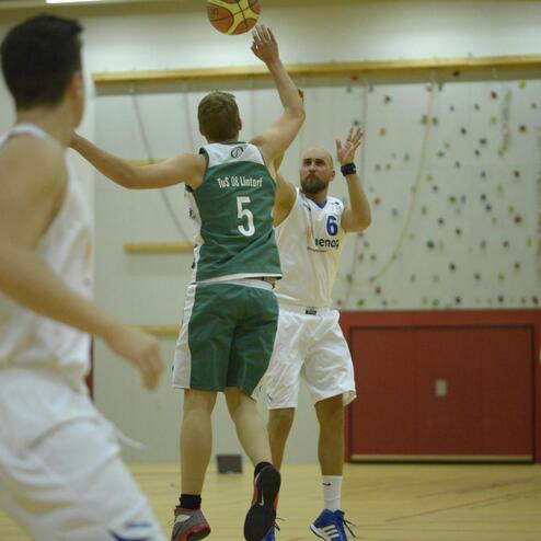 Basketballspiel in der Halle: Ein Spieler mit Ball springt zum Wurf, während andere Spieler in Position stehen.
