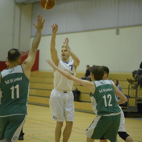 Basketballspieler in weißer Kleidung wirft Ball über zwei Verteidiger in grüner Kleidung auf einem Basketballfeld.