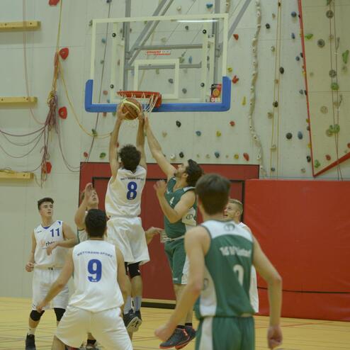 Spielszene eines Basketballspiels: Spieler in weiß springt, um den Ball im Korb zu versenken, während Mitspieler zuschauen.