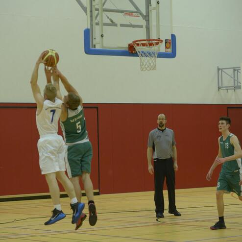 Zwei Basketballspieler springen zum Wurf, während ein Schiedsrichter das Geschehen beobachtet.