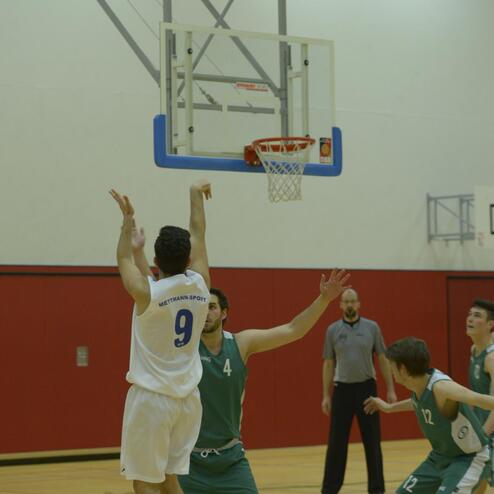 Basketballspieler im weißen Trikot springt zum Wurf, während Gegner in grünen Trikots ihn zu blockieren versuchen.