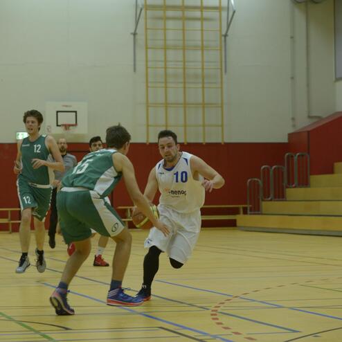 Zwei Basketballspieler in Aktion auf dem Spielfeld; einer in weißem Trikot dribbelt den Ball, der andere in grünem Trikot verteidigt.