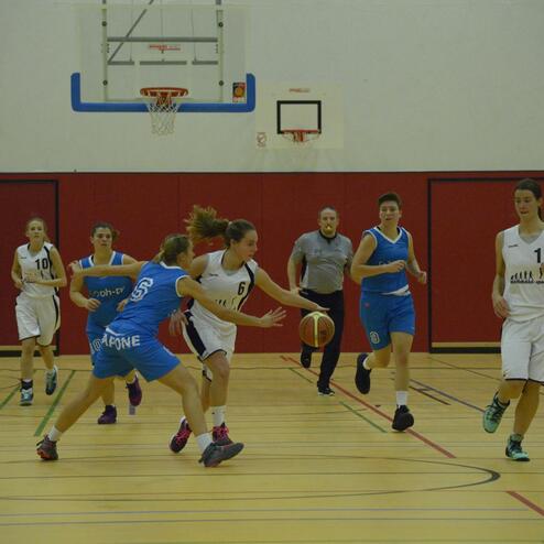 Eine Basketballspielerin in Weiß dribbelt einen Ball, während Spielerinnen in Blau sie verfolgen.