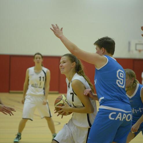 Zwei Basketballmannschaften befinden sich im Wettkampf auf dem Spielfeld, eine Spielerin hält den Ball.