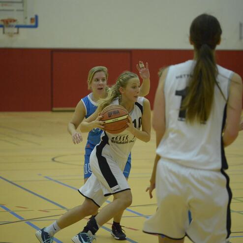 Basketballspiel: Spielerin mit Ball dribbelt in Richtung Korb, während Gegnerin versucht, den Ball zu ergattern.
