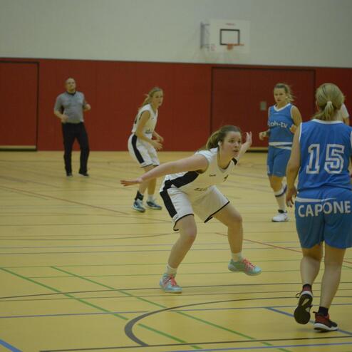 Zwei Basketballspielerinnen auf dem Feld, eine verteidigt, während die andere mit dem Ball dribbelt. Zuschauer im Hintergrund.
