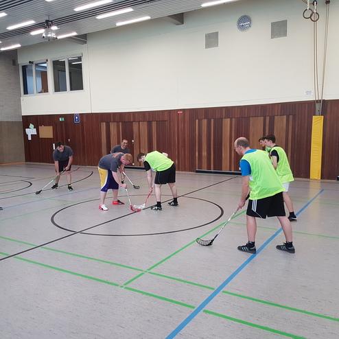 Gruppenübung in einer Sporthalle mit acht Personen, die Floorball spielen, einige in Neonwesten.