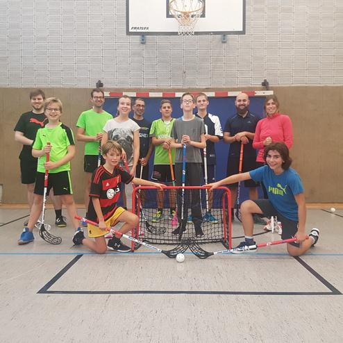 Gruppenshot von Jugendlichen und Erwachsenen mit Floorball-Schlägern vor einem Tor in einer Sporthalle.