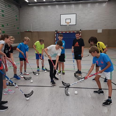 Gruppe von Kindern in Sportkleidung übt mit Hockeyschlägern und Bällen in einer Turnhalle.