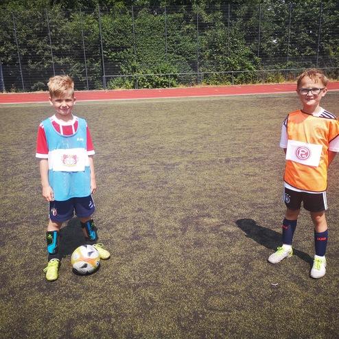 Zwei Kinder in Fußballtrikots stehen auf einem Kunstrasenplatz, ein Ball liegt zwischen ihnen.