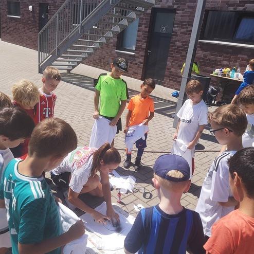 Gruppe von Kindern mit T-Shirts, die um eine Person versammelt sind, die auf den Boden zeichnet. Sonniger Tag im Freien.