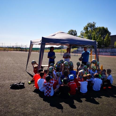 Kinder sitzen unter einem Zelt auf einem Sportplatz, während Trainer ihnen Fußballtechniken mit Bällen demonstrieren.