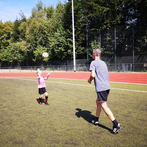 Ein Kind in pinkem Trikot fängt einen Fußball, während ein Erwachsener auf einem Sportplatz mit Laufbahn steht.