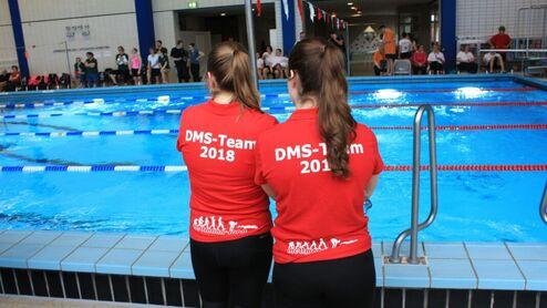 Zwei Personen mit roten T-Shirts stehen am Schwimmbecken und beobachten den Wettkampf. Auf den T-Shirts steht „DMS-Team 2018“.