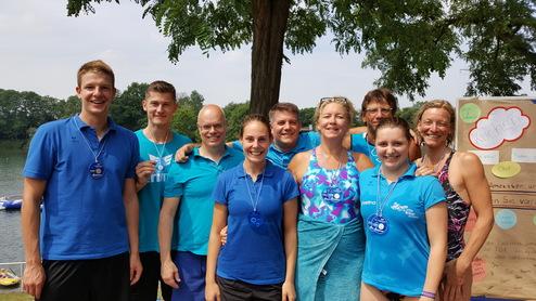 Gruppenfoto von neun Menschen in blauen Shirts mit Medaillen, vor einem See und einem Plakat im Hintergrund.