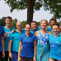 Gruppenfoto von neun Menschen in blauen Shirts mit Medaillen, vor einem See und einem Plakat im Hintergrund.