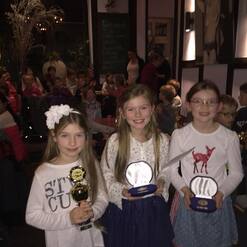 Drei Mädchen halten Pokale und Medaillen in einem festlich dekorierten Raum voller Zuschauer.