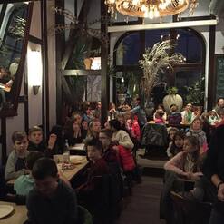 Menschen sitzen in einem gemütlichen Restaurant, viele Kinder, einige Erwachsene, im Hintergrund Pflanzen und Dekoration.