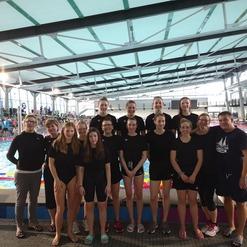 Gruppenfoto von Schwimmerinnen in einheitlichen schwarzen Badeanzügen vor einem Schwimmbecken in einem modernen Hallenbad.