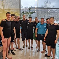 Gruppe von acht jungen Männern in Schwimmbekleidung lächelt am Beckenrand eines Schwimmbeckens.